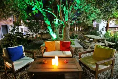 Bozcaada Alize Otel’in gece ışıklarıyla aydınlanan bahçesi, zeytin ağacı altında renkli minderli koltuklar ve sıcak bir ada akşamı atmosferi.